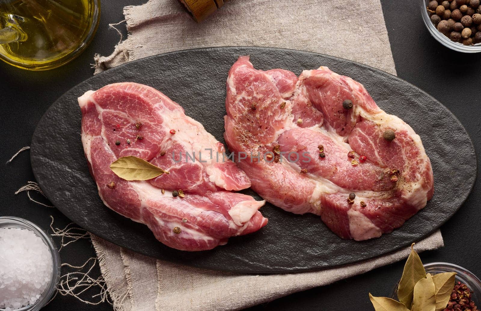 Two raw pork neck steaks on a board and spices for cooking. Top view of black table