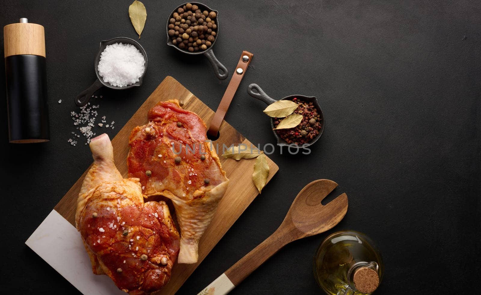 Raw chicken drumsticks seasoned on a wooden board, accompanied by salt and peppercorns, viewed from the top. Black table