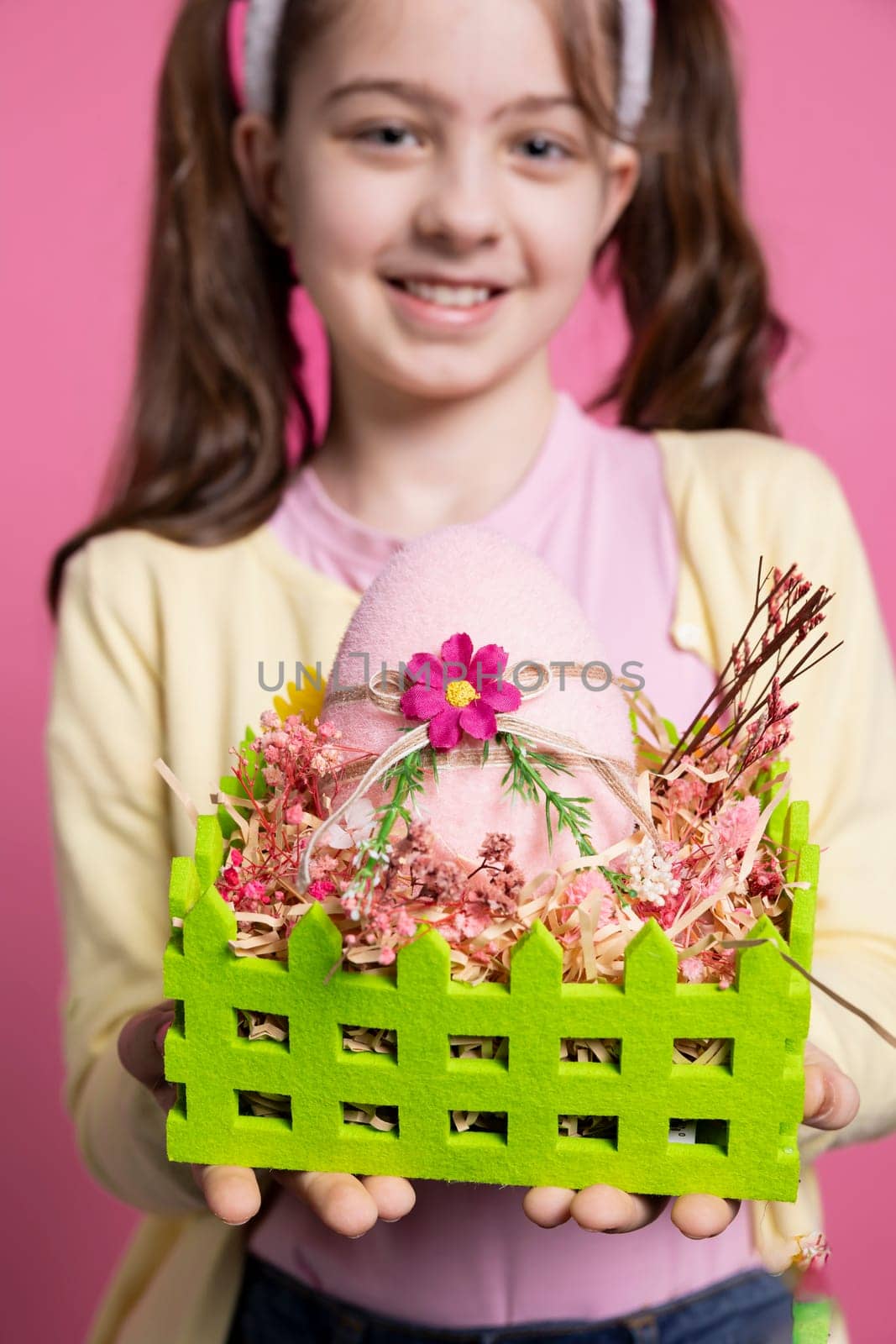 Positive excited small kid showing her handmade easter eggs in a colored basket, feeling proud of her arrangement. Young child with bunny ears holding festive decorations, april celebrations.