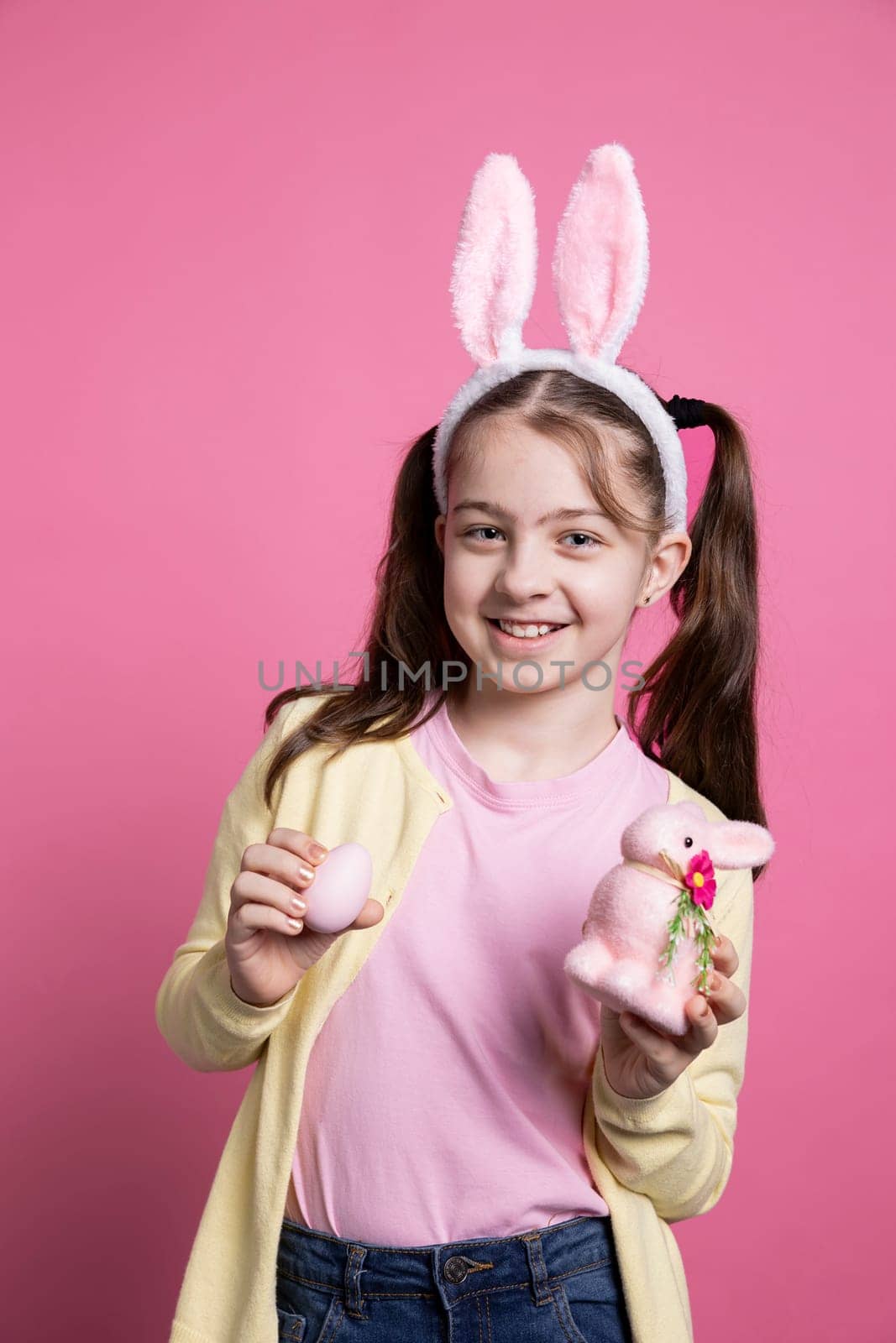 Optimistic small kid posing with painted eggs and a pink rabbit toy, wearing fluffy bunny ears and being cheerful in front of camera. Young girl with pigtails showing her easter decorations.