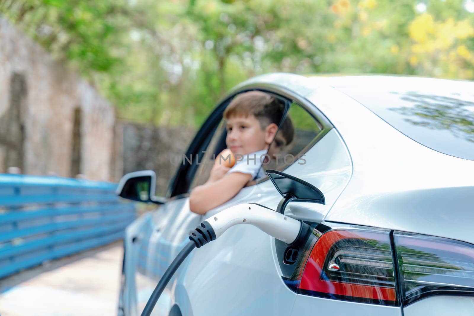 EV car recharge battery from EV charging station with blur little boy waiting inside the car in background. Road trip travel by alternative vehicle powered by clean and sustainable energy. Perpetual