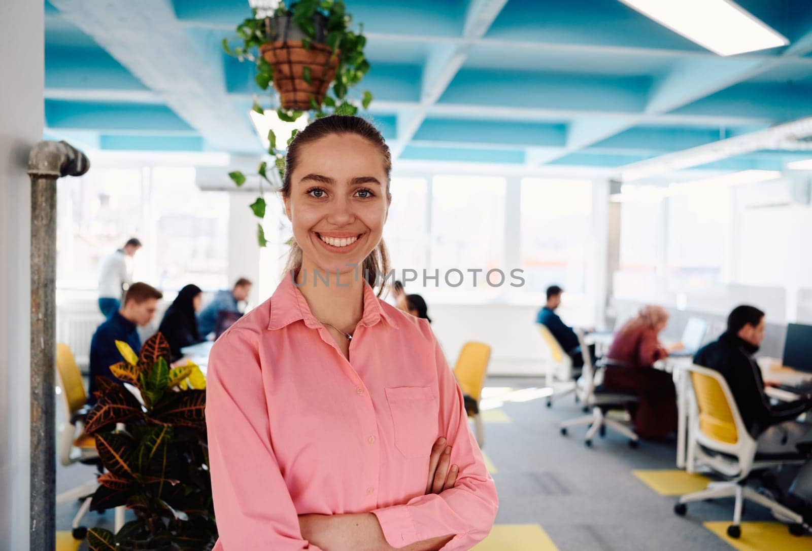 Portrait of young smiling business woman in creative open space coworking startup office. Successful businesswoman standing in office with copyspace. Coworkers working in background.