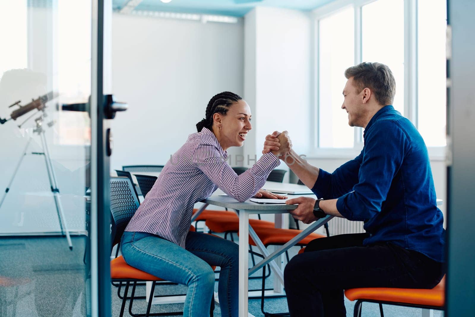 In a modern startup office, a businessman and a businesswoman business colleagues engage in a symbolic arm-wrestling match, reflecting teamwork, competition, and innovation in their dynamic workplace