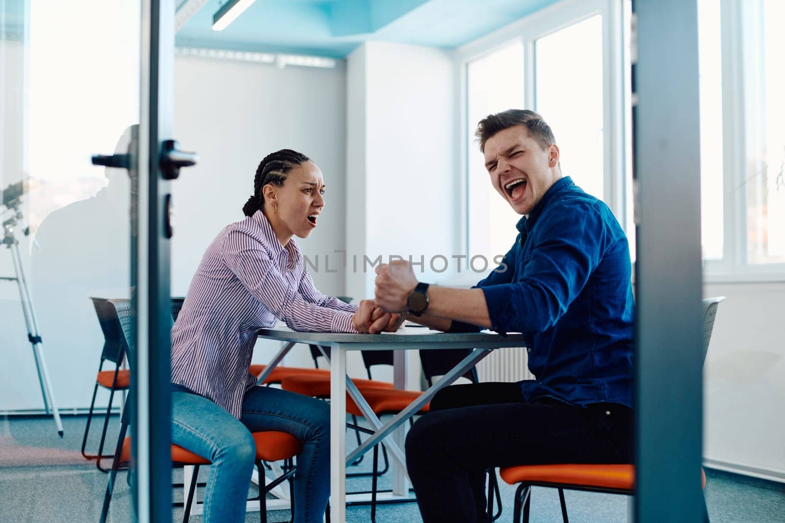 In a modern startup office, a businessman and a businesswoman business colleagues engage in a symbolic arm-wrestling match, reflecting teamwork, competition, and innovation in their dynamic workplace. by dotshock
