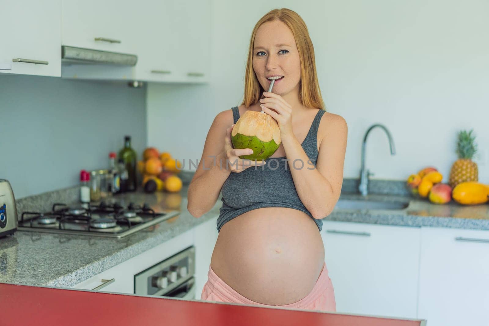 Quenching her pregnancy thirst with a refreshing choice, a pregnant woman joyfully drinks coconut water from a coconut in the kitchen, embracing natural hydration during this special journey by galitskaya