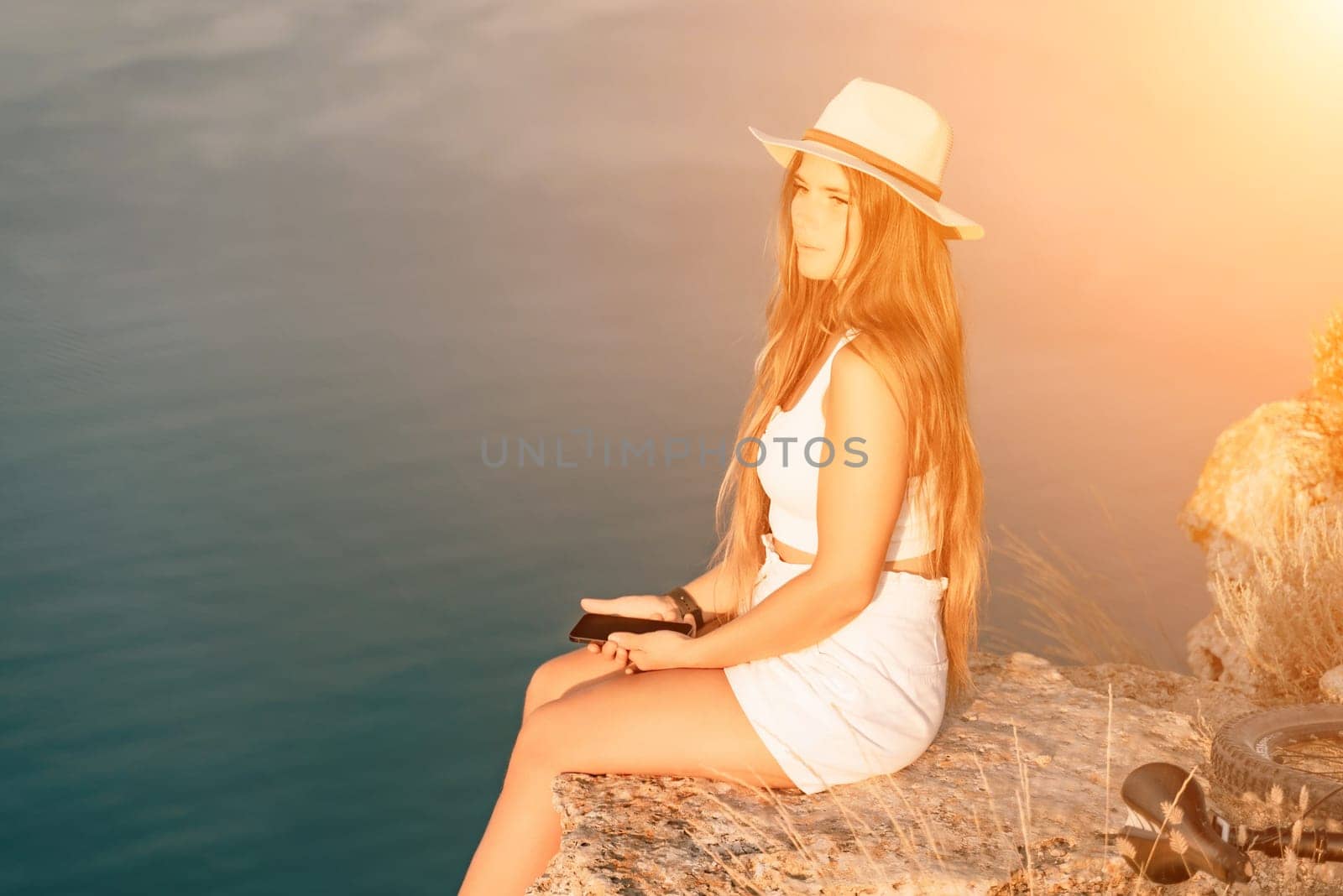 A tourist woman is sitting by the sea in a hat and white summer clothes, looking happy and relaxed