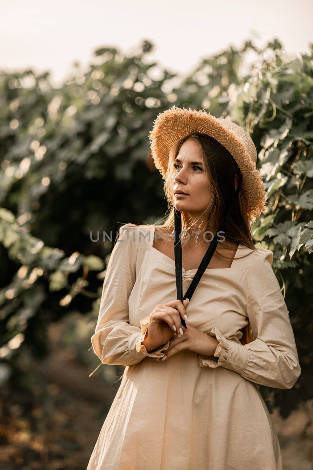 Woman with straw hat stands in front of vineyard. She is wearing a light dress and posing for a photo. Travel concept to different countries.