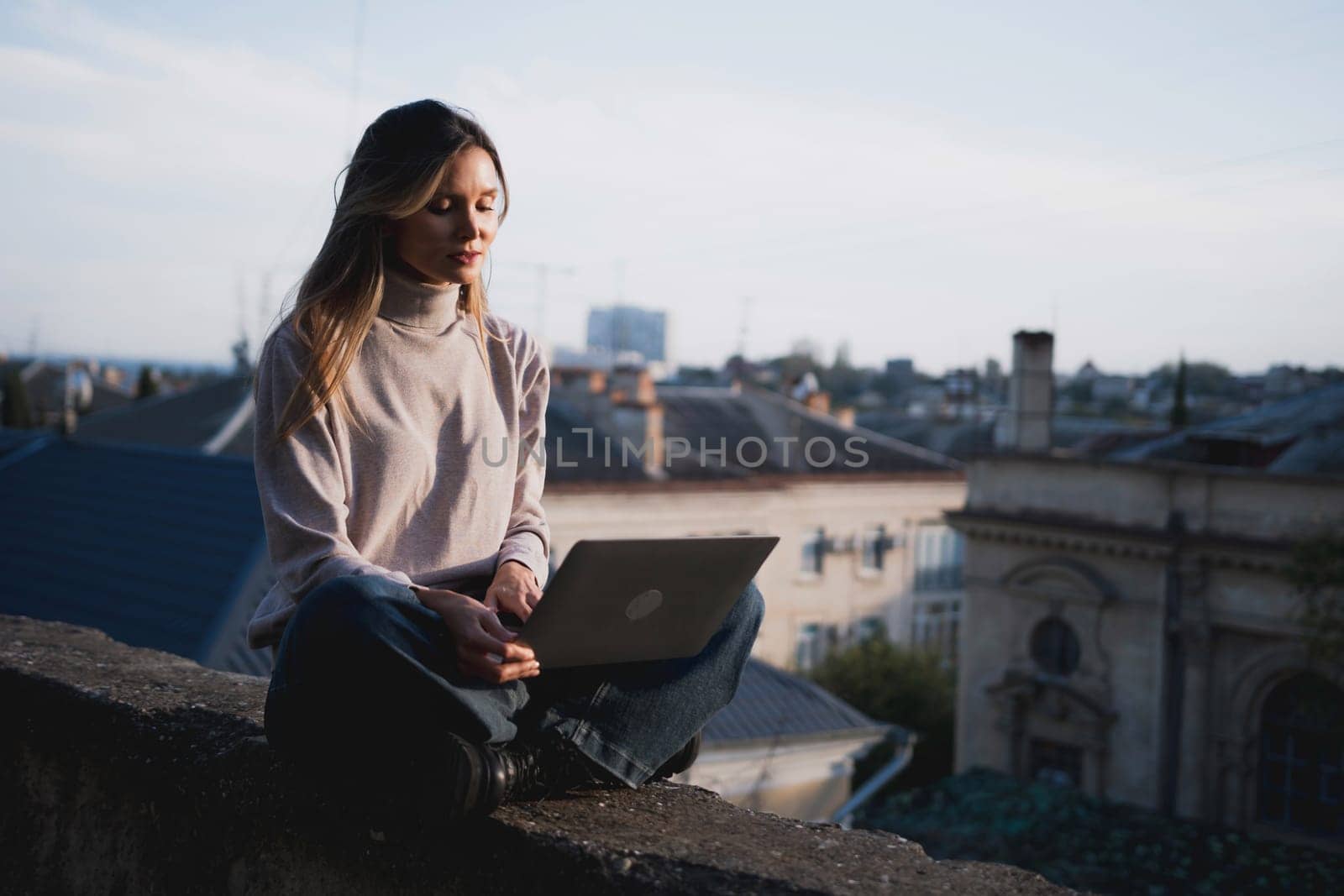 Woman freelancer uses laptop on cement wall outdoors against the sky and the roof of the city. The woman to be focused on her work or enjoying some leisure time while using her laptop