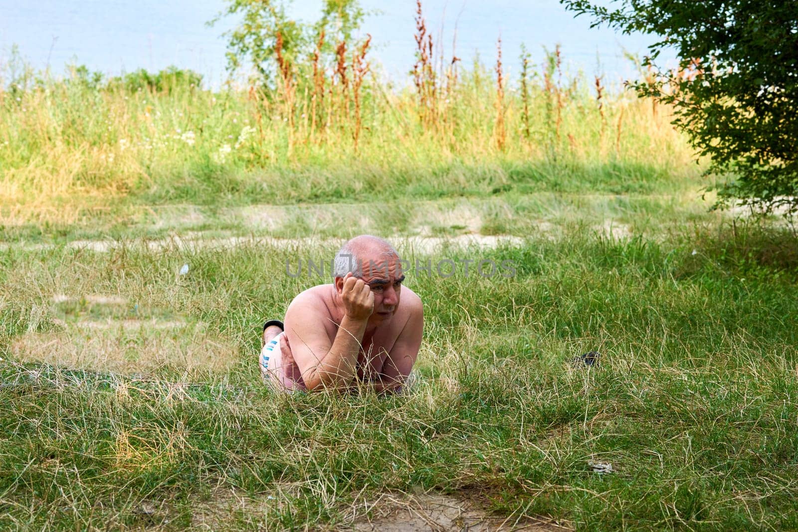 An elderly man is resting on a grass in a lawn near a summer river by jovani68