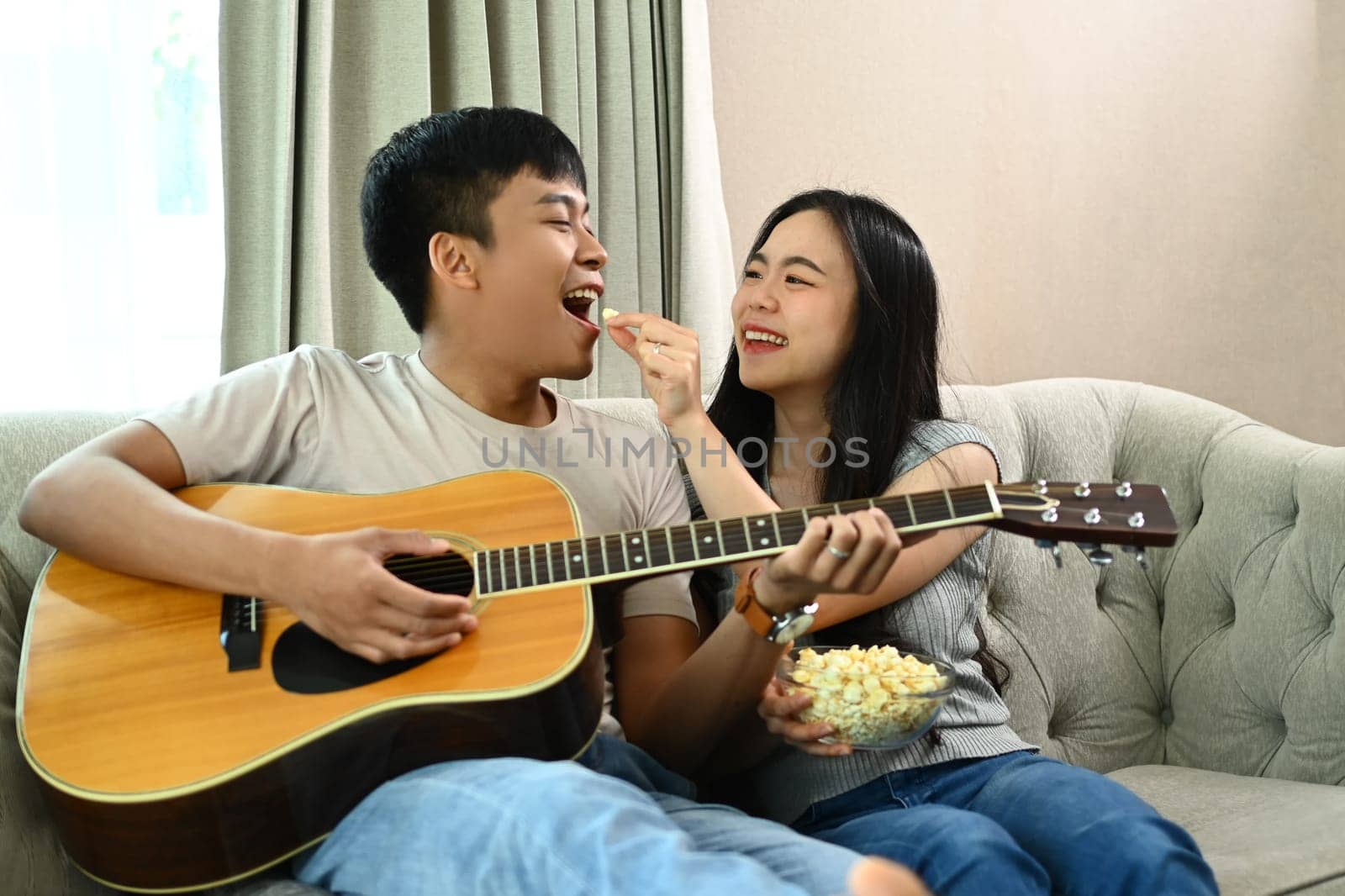 Happy man playing guitar while his wife feeding him with popcorns, enjoy weekend activity together by prathanchorruangsak