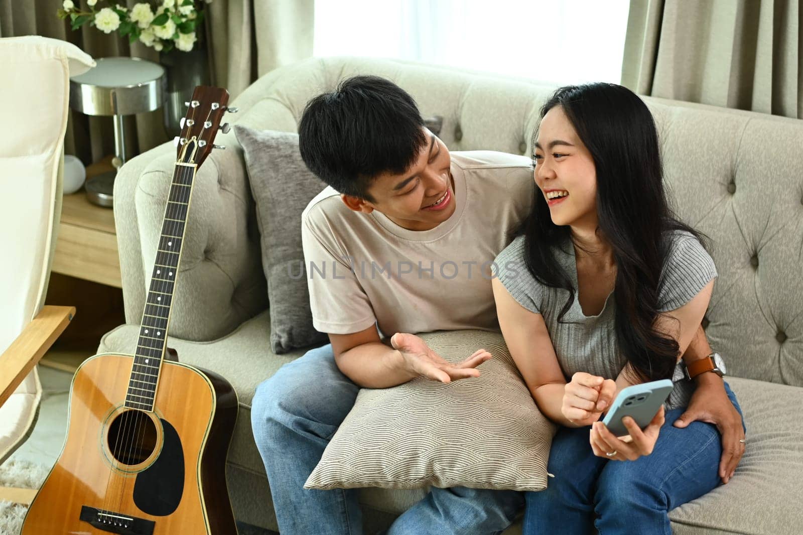 Cheerful young Asian couple using smartphone on comfortable couch in living room by prathanchorruangsak