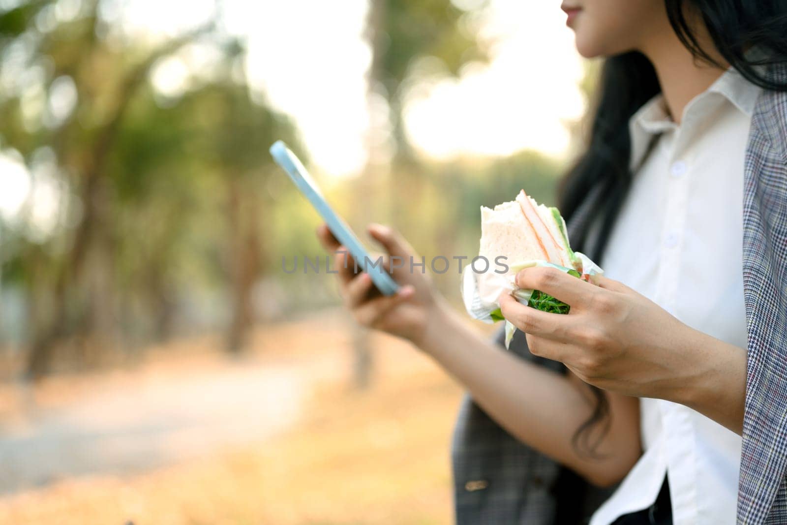 Cropped shot young female office worker having lunch in the park and using smartphone by prathanchorruangsak