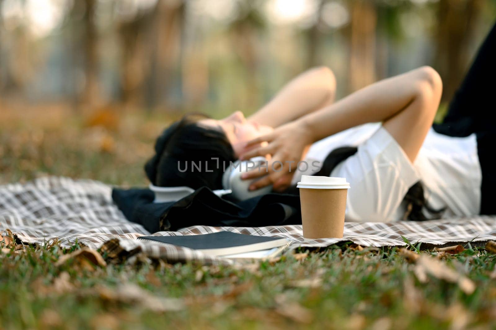 A paper cup of coffee on checkered blanket near relaxed young woman listening to music with headphone by prathanchorruangsak