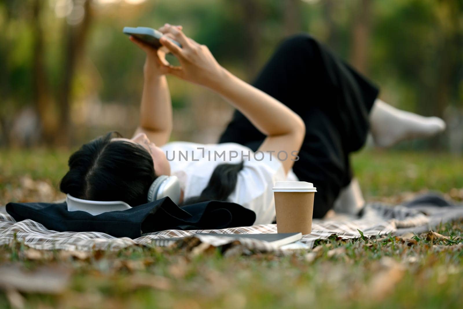 Carefree young woman lying on blanket in the park and using mobile phone.