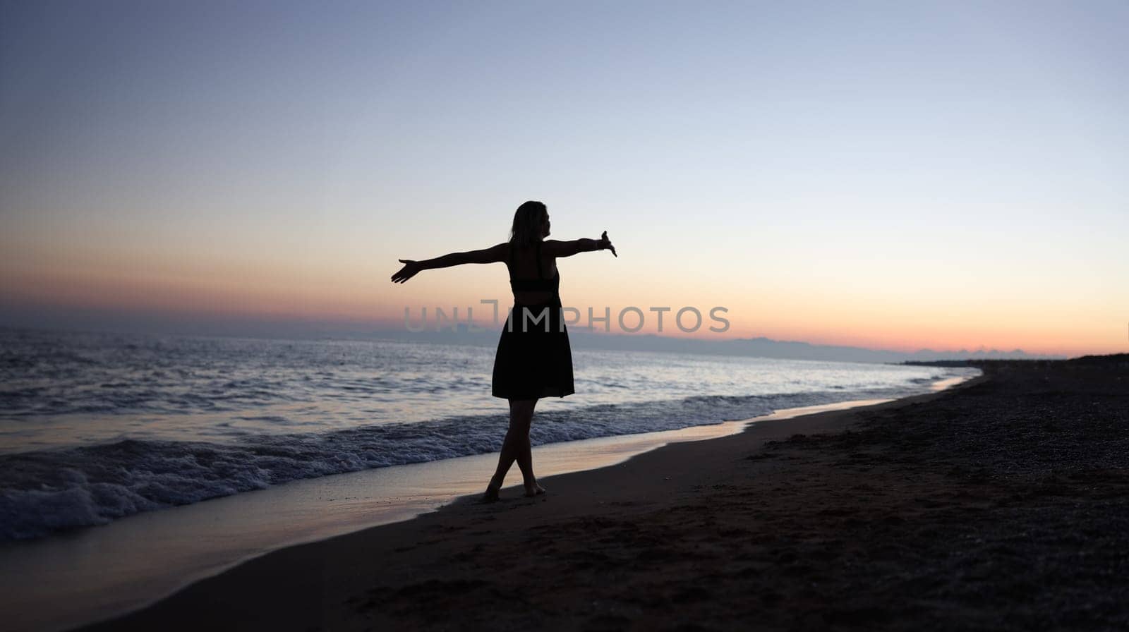 Woman standing with open arms on seashore at sunset by kuprevich