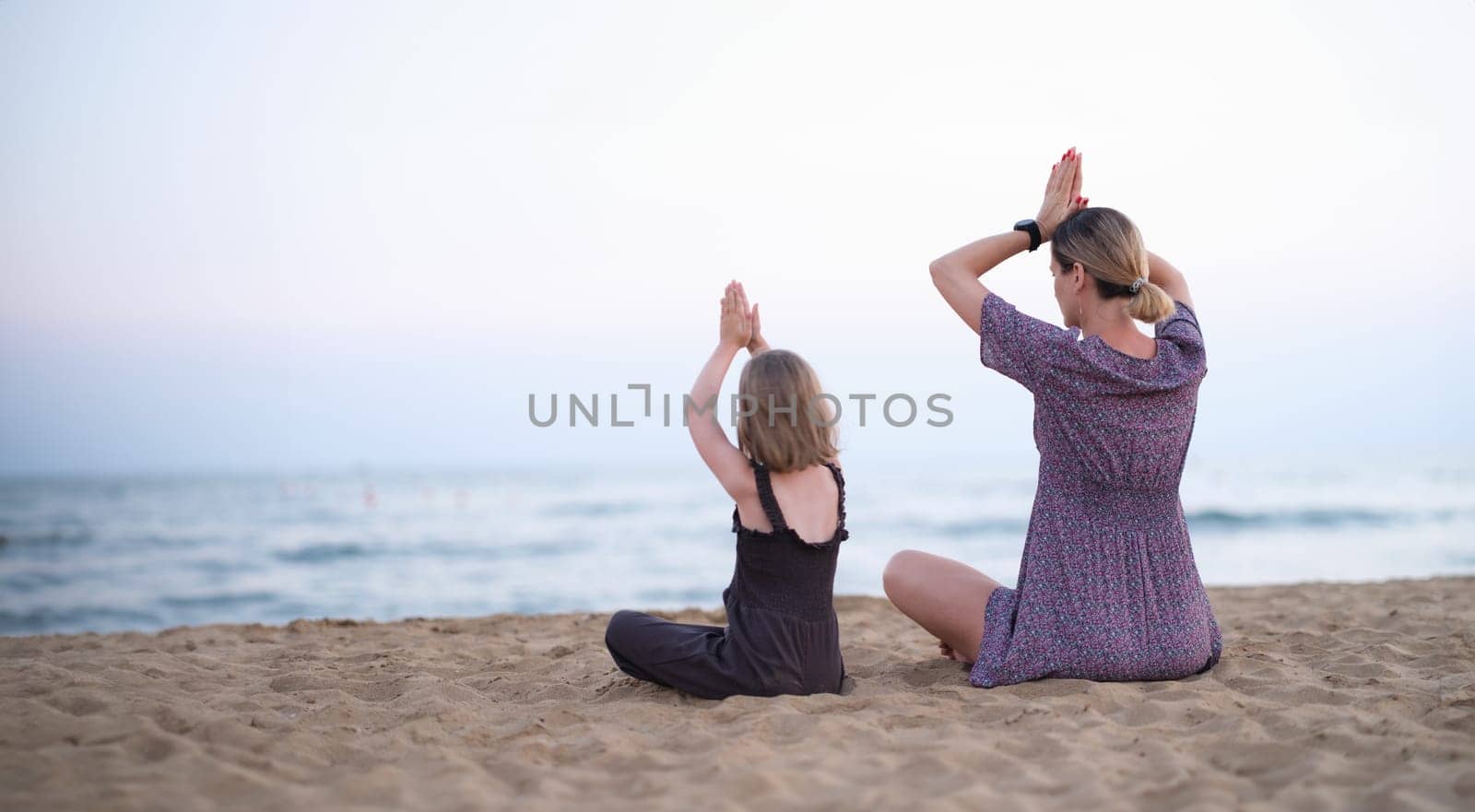 Happy family mother and daughter do yoga, meditate in lotus position on beach by kuprevich