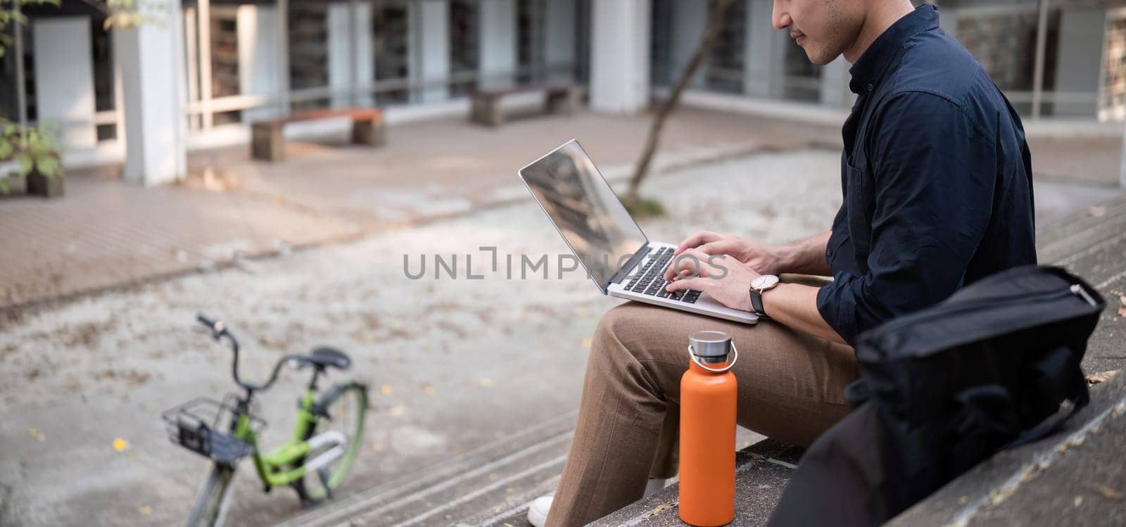 A young Asian businessman rides a bicycle to work. Sitting outside the office working using a laptop..