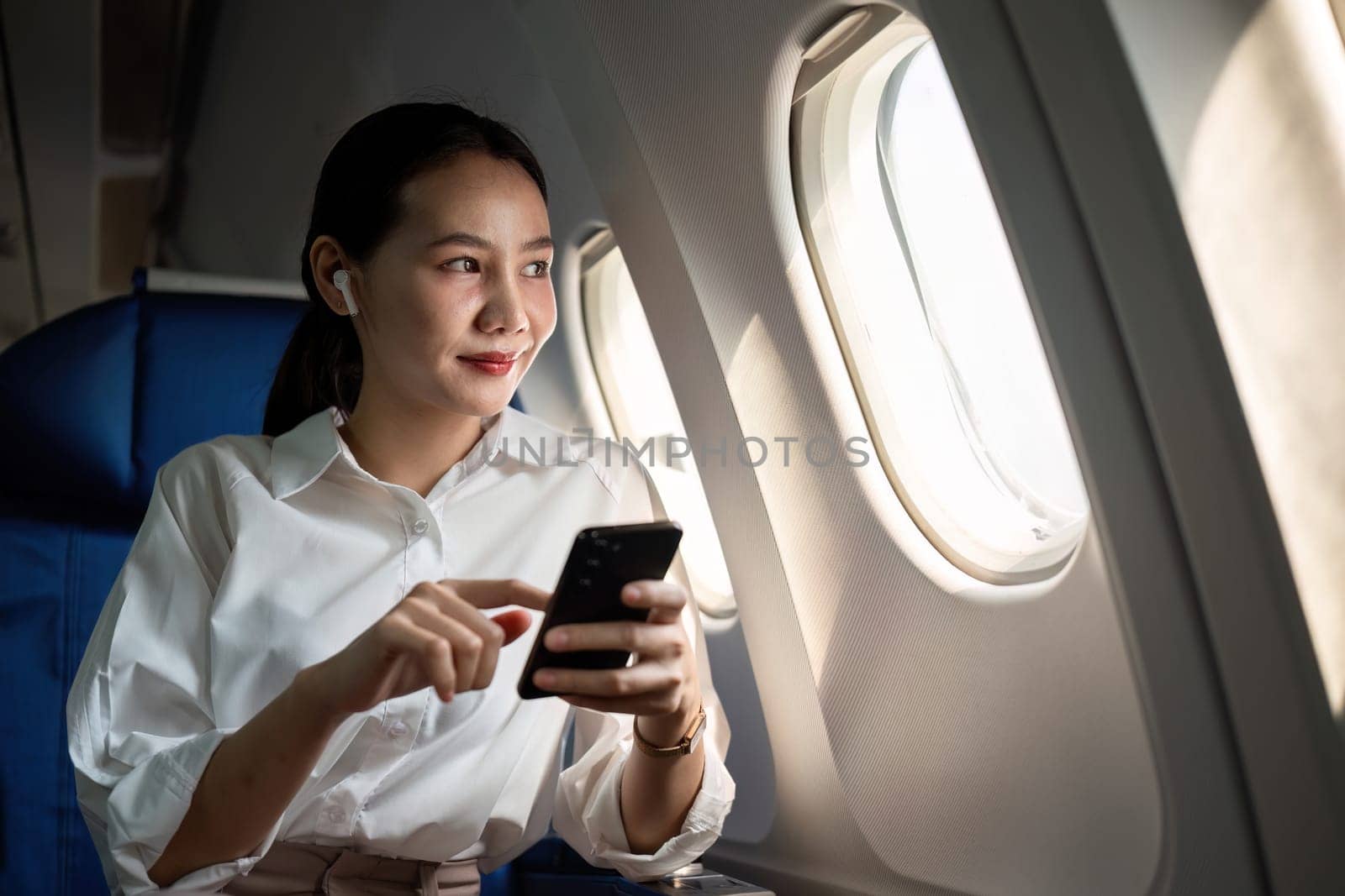 Young Asian woman checks business news on mobile phone, sitting near window in first class on airplane during flight, travel and business concept by wichayada