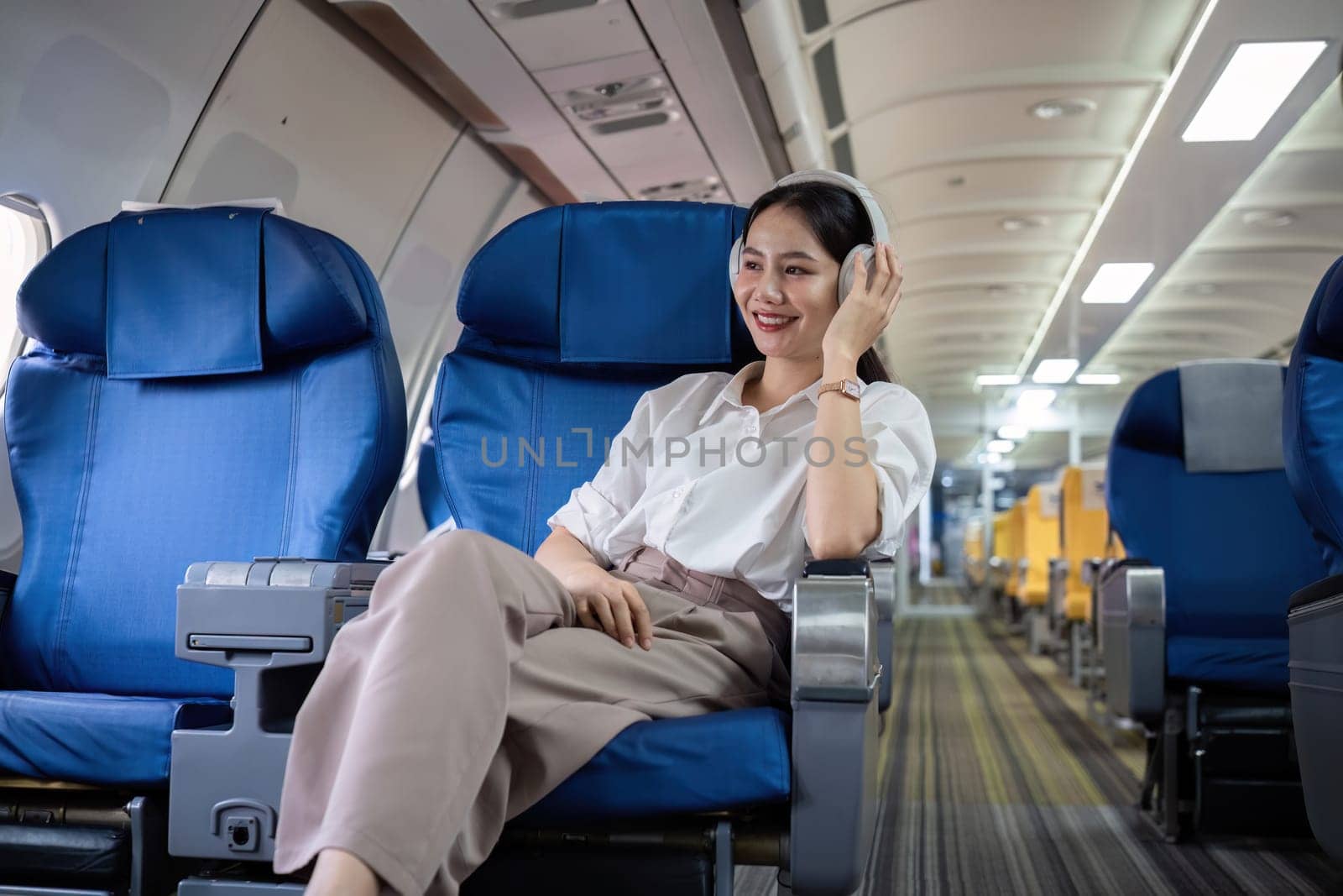 Young woman wearing headphones listening to music during travel, sitting near window in first class on airplane during flight, travel and business concept.