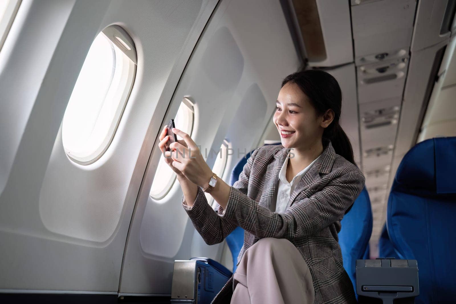 Young female traveler sits near the window in first class on a plane during a flight. Use your cell phone to take photos of the view outside your window, travel, and business ideas. by wichayada