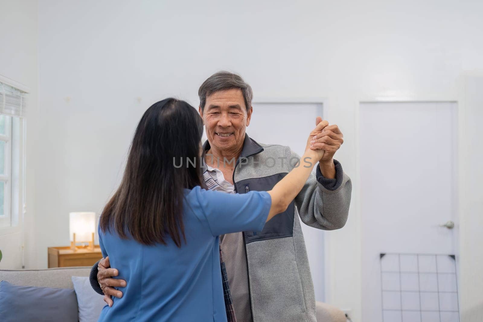 Elderly Asian couple in their 60s dancing in the living room together, enjoying music and smiling in love..