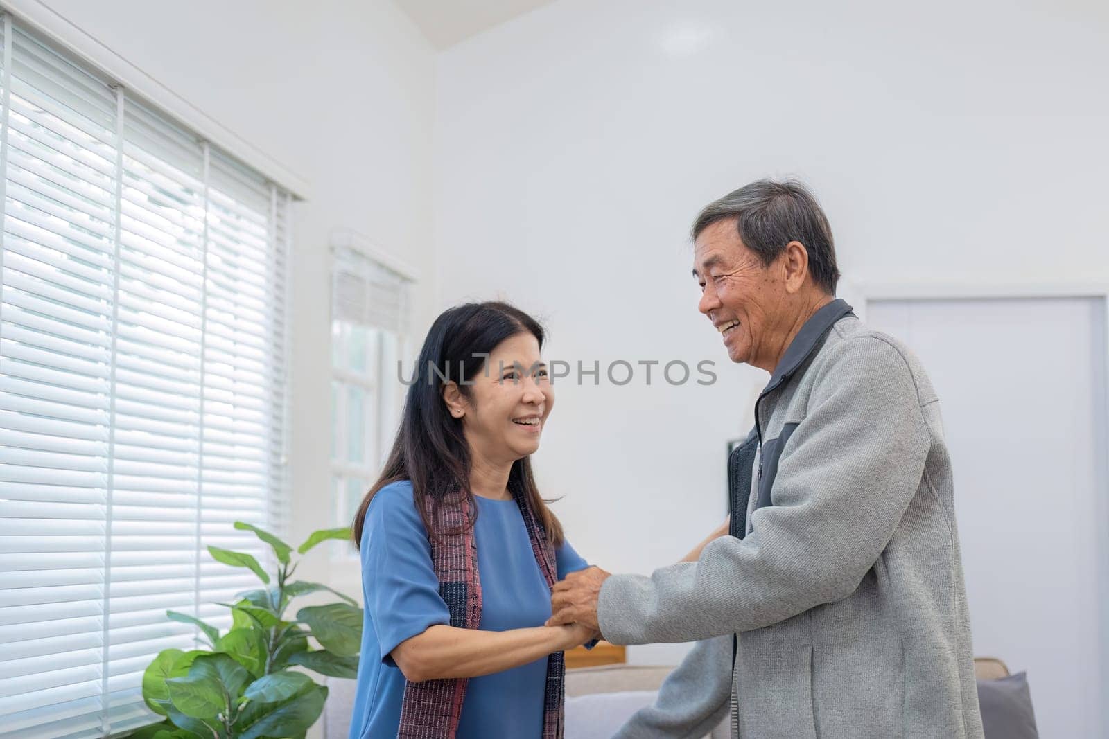 Elderly Asian couple in their 60s dancing in the living room together, enjoying music and smiling in love..