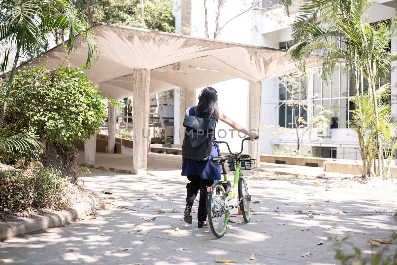 A young Asian woman commutes to work by cycling in a green city, carrying a backpack and using a reusable drinking cup to avoid harming the environment. by wichayada