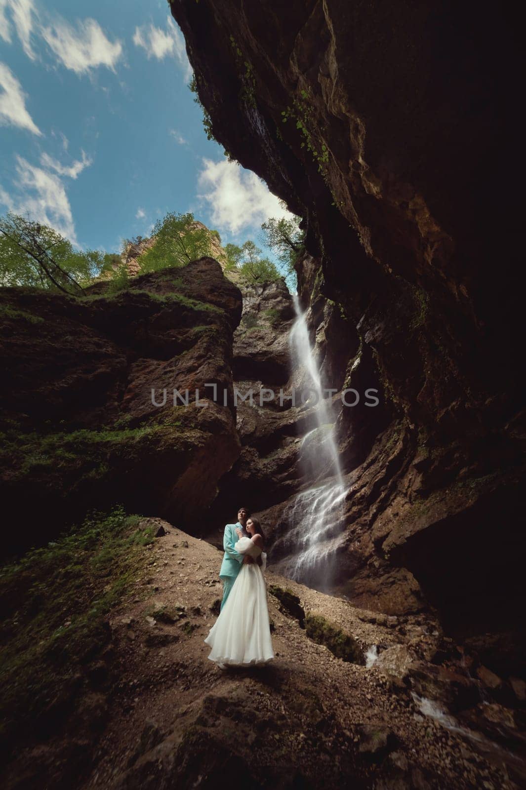 A young man and his wife are standing in an embrace in a canyon against the backdrop of a high waterfall. Newlyweds wedding couple in the mountains.