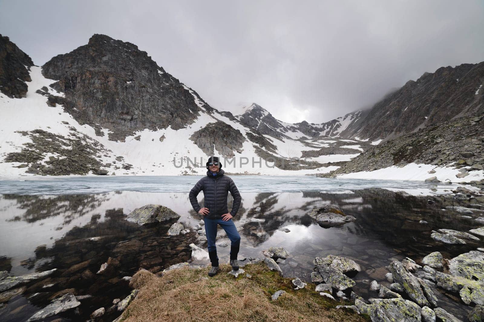 Happy active cheerful tourist in mountain sunglasses and a down jacket, joyful smiles expressively rejoices against the backdrop of a mountain lake high in the mountains.