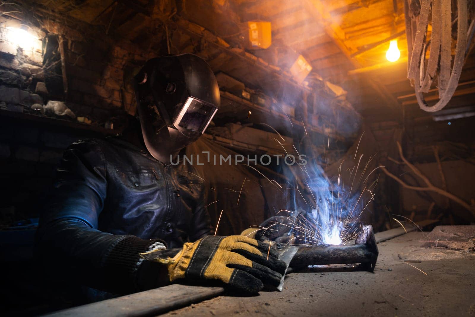 Young mechanic with a welding machine in an old dirty garage at night. Man in protection doing welding work, hobby by yanik88