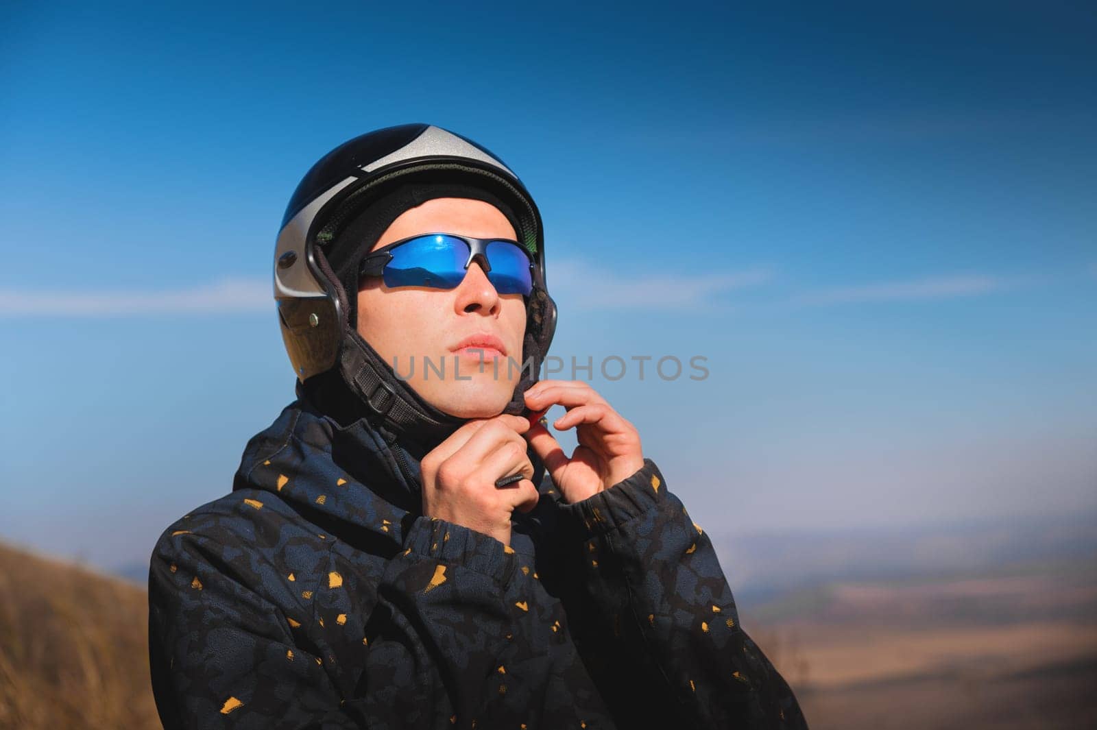 A young male paraglider in sunglasses fastens his helmet on a sunny day. Preparing for paragliding.