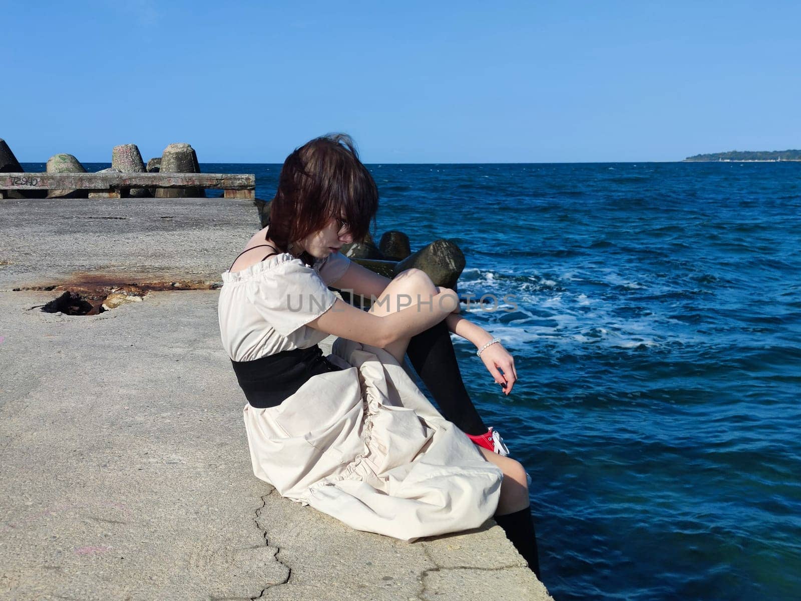 sad teenage girl in a white dress sitting on an old sea pier.