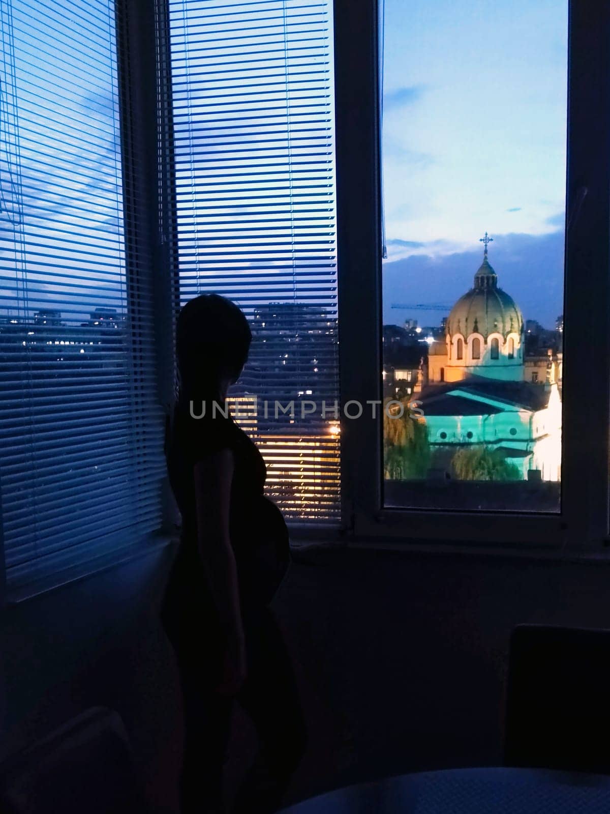 a pregnant woman thoughtfully looks out the window at the cathedral in the evening.