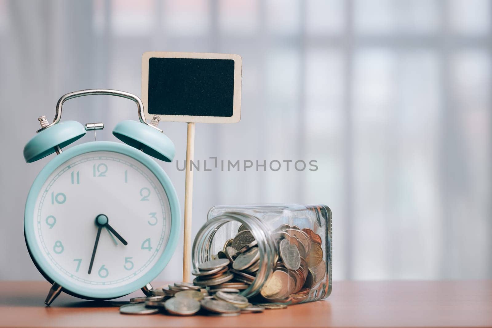 Alarm clock and coins on a wooden table, accompanied by a blank chalkboard for messages or notes, symbolizing the integration of time management and financial planning.