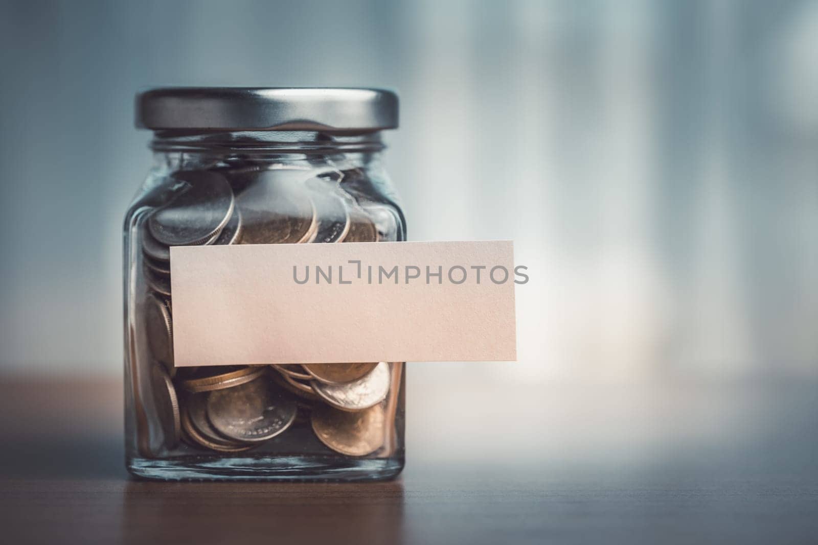 Coins in a glass jar with blank paper label on wooden table by iamnoonmai