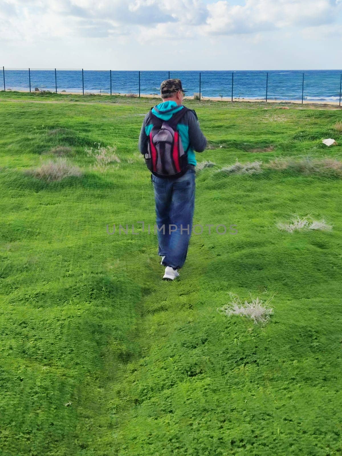 a man with a backpack walks along the green grass towards the sea, rear view by Annado