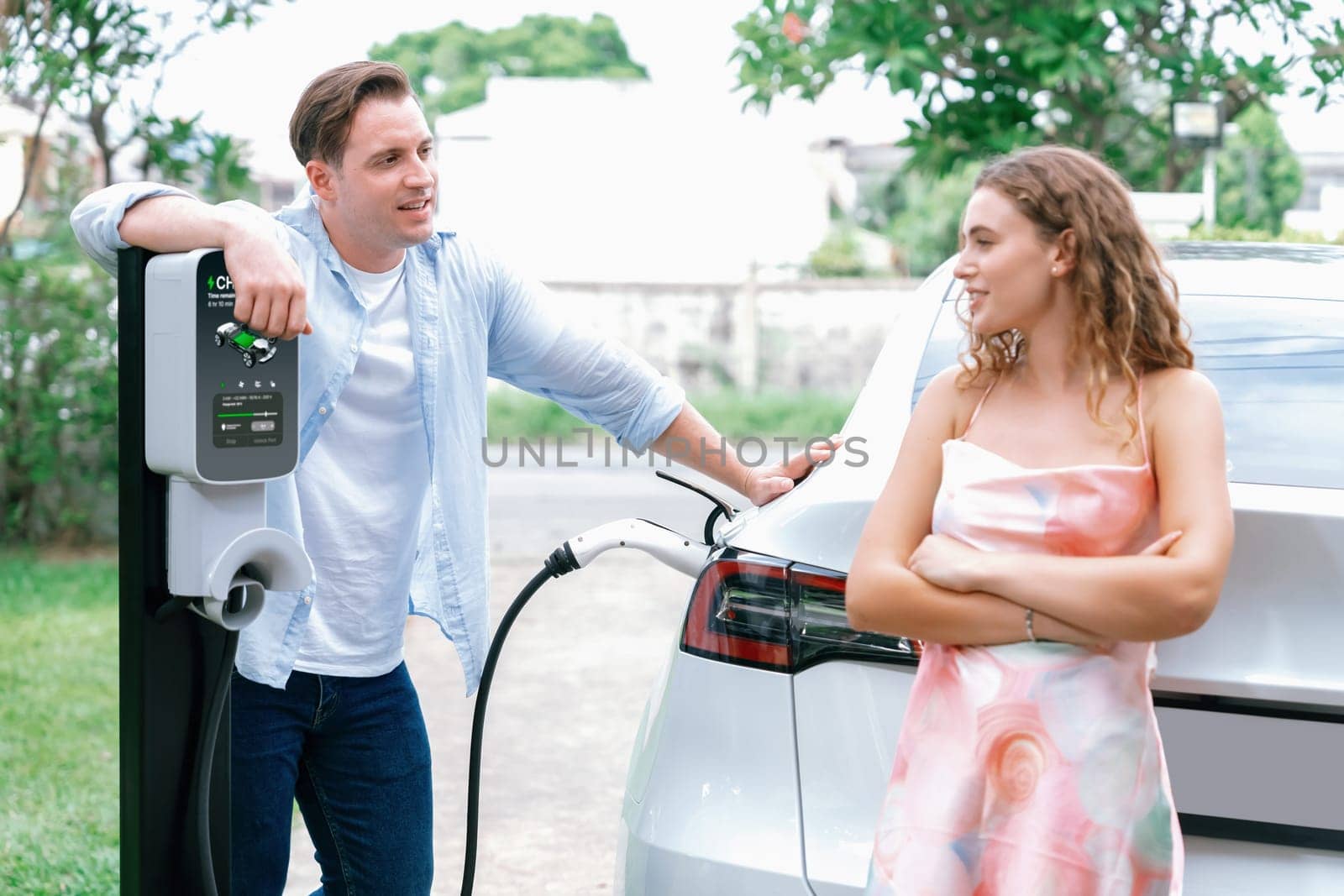 Happy and lovely couple with eco-friendly conscious recharging electric vehicle from EV charging station. EV car technology utilized as alternative transportation for future sustainability. Synchronos