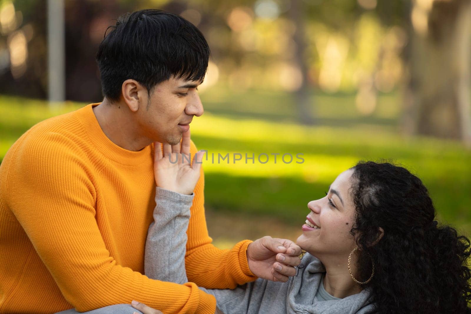 happy latin woman touching face of boyfriend and sitting on grass with copy space by papatonic