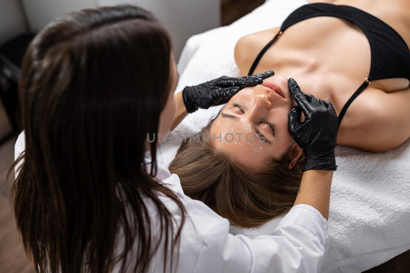 Beautiful young woman in black bikini getting face lifting massage in cosmetology clinic.
