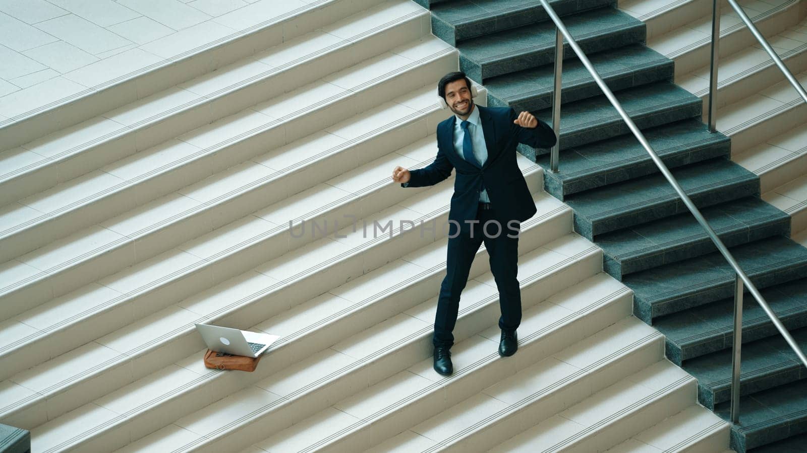 Professional business man standing at stair while moving to music from headphone. Skilled project manager dancing while listening relax sound with laptop placed on stairs. Modern dance. Exultant.