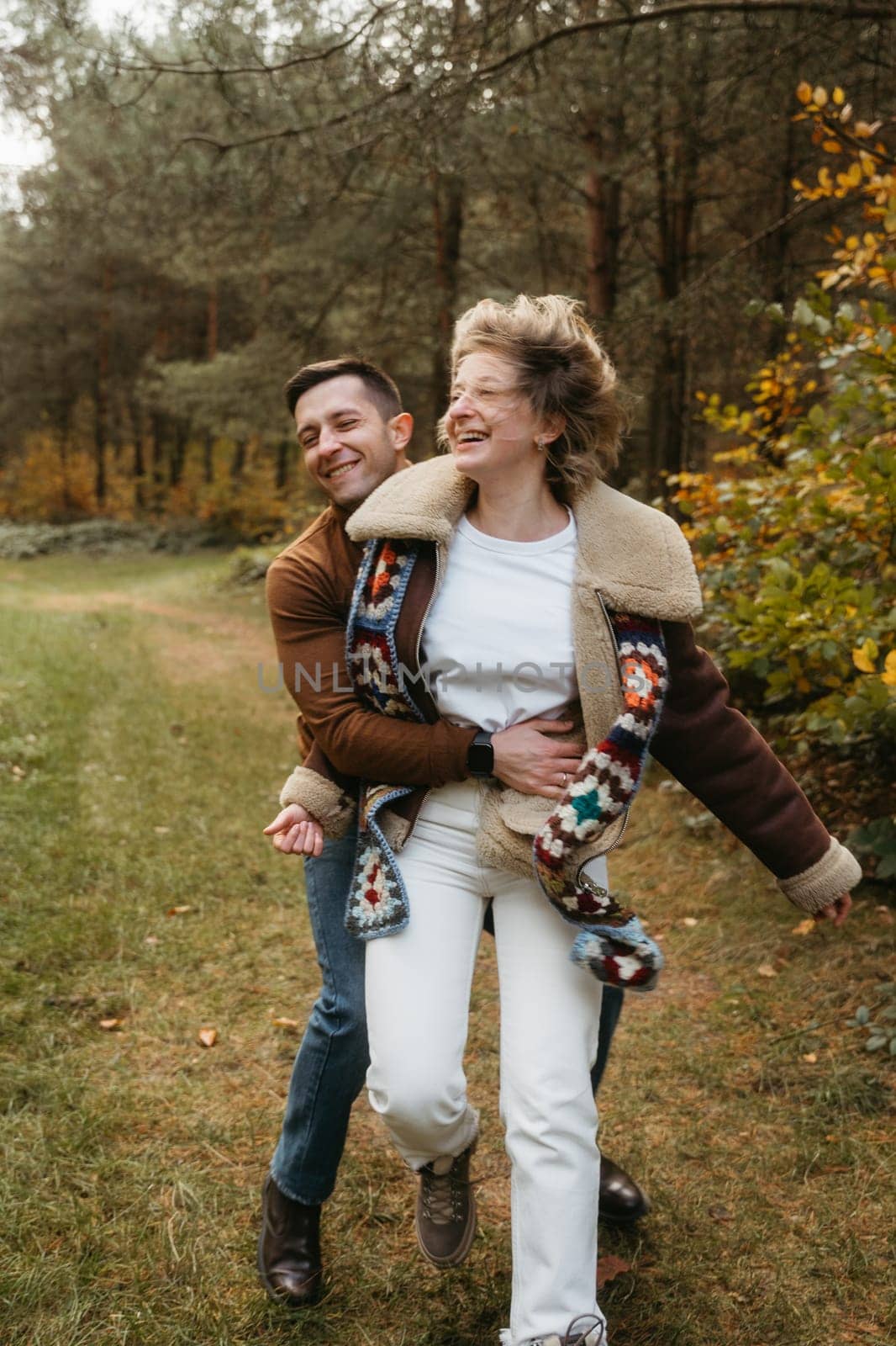 Happy couple is having fun in the middle of the forest, a smiling man hugging his girlfriend in an autumn park. by Romvy