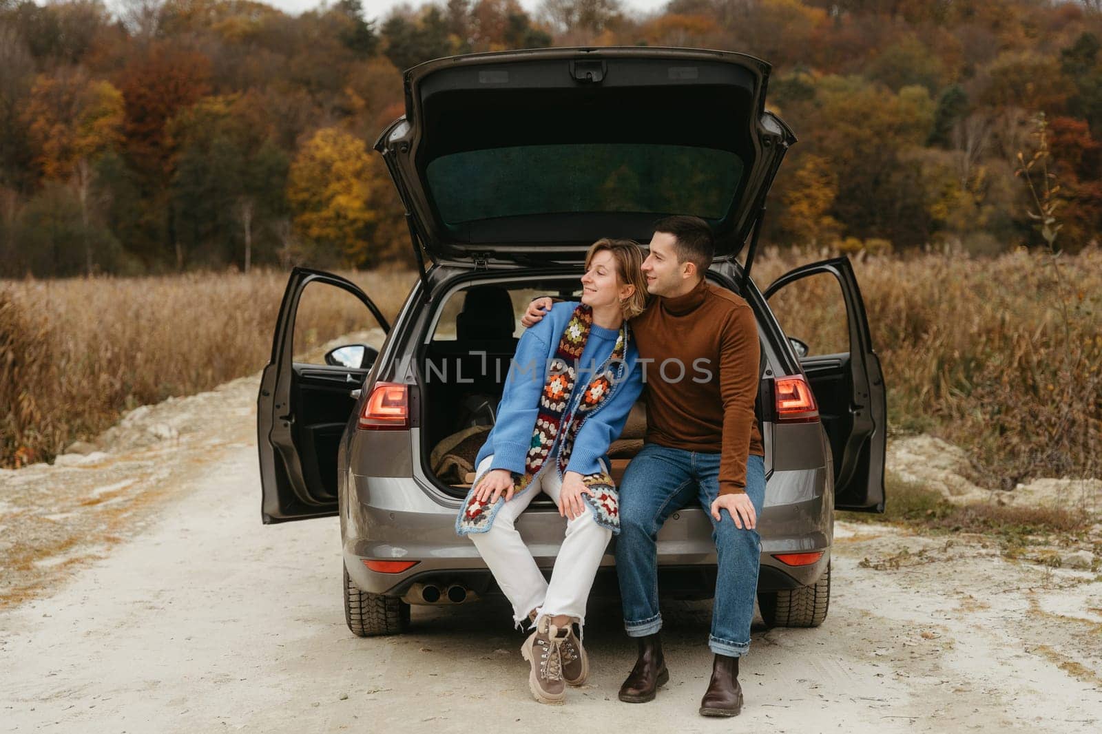 Happy young woman and man sitting in the open trunk of their car while traveling in autumn and looking away, couple road trip concept by Romvy