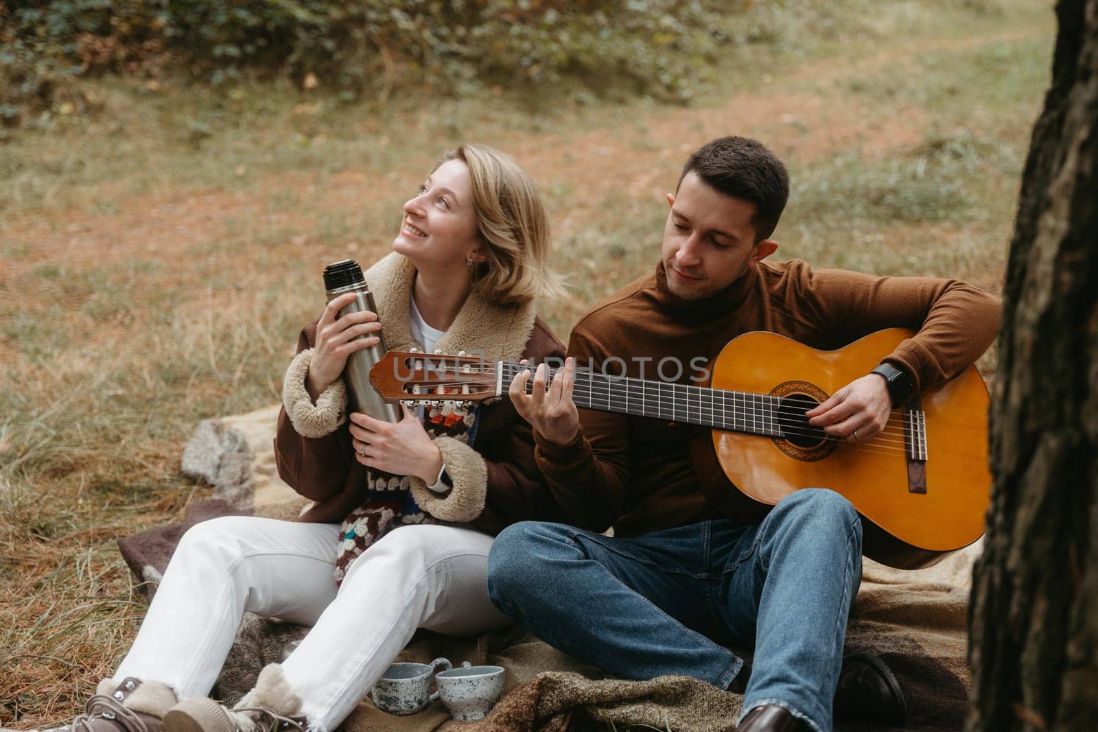 Adult couple sitting on blanket in autumn park, a man playing guitar for happy woman