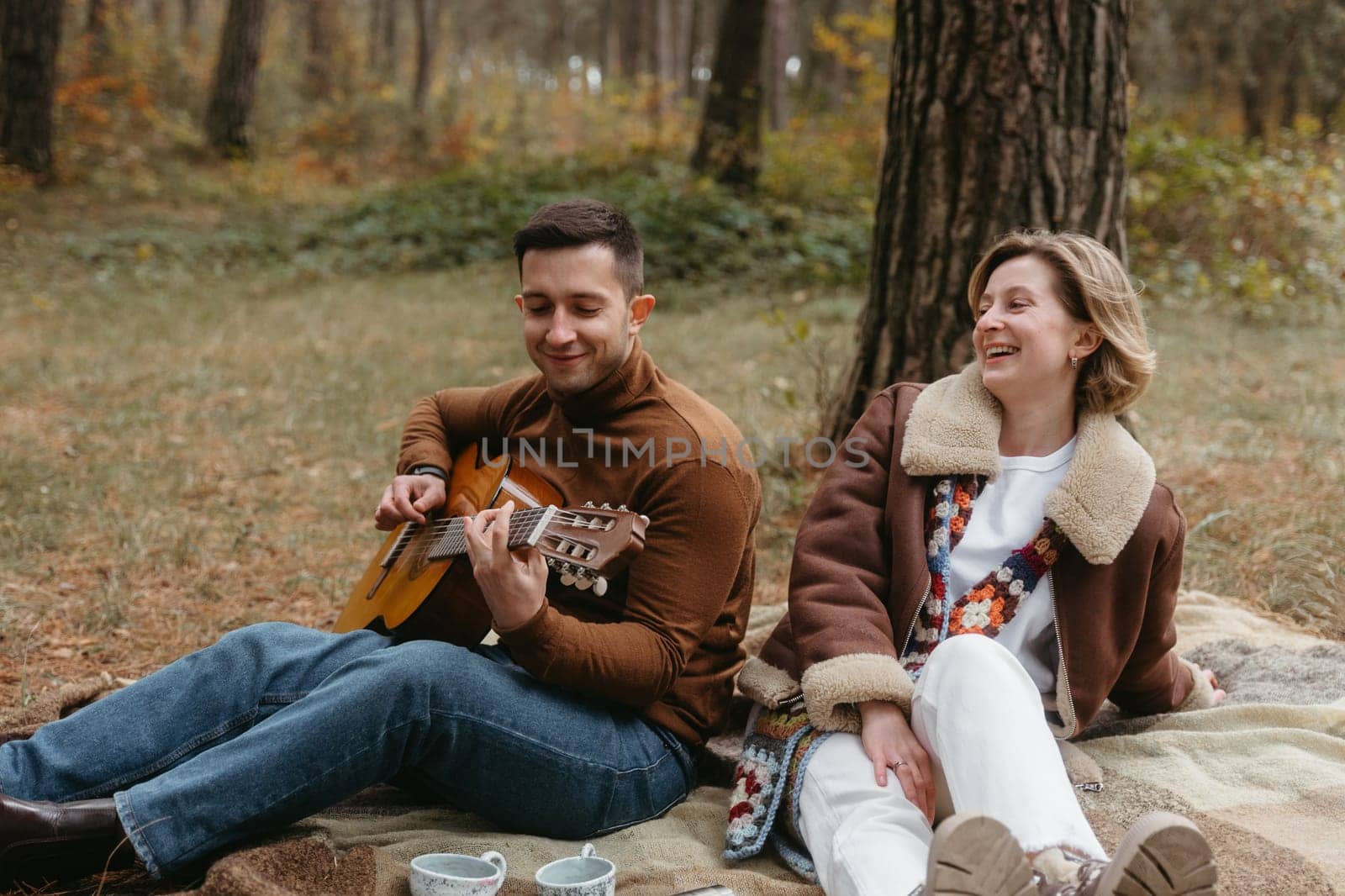 A man is playing a guitar for a happy woman while they are sitting on a blanket in the autumn woods by Romvy