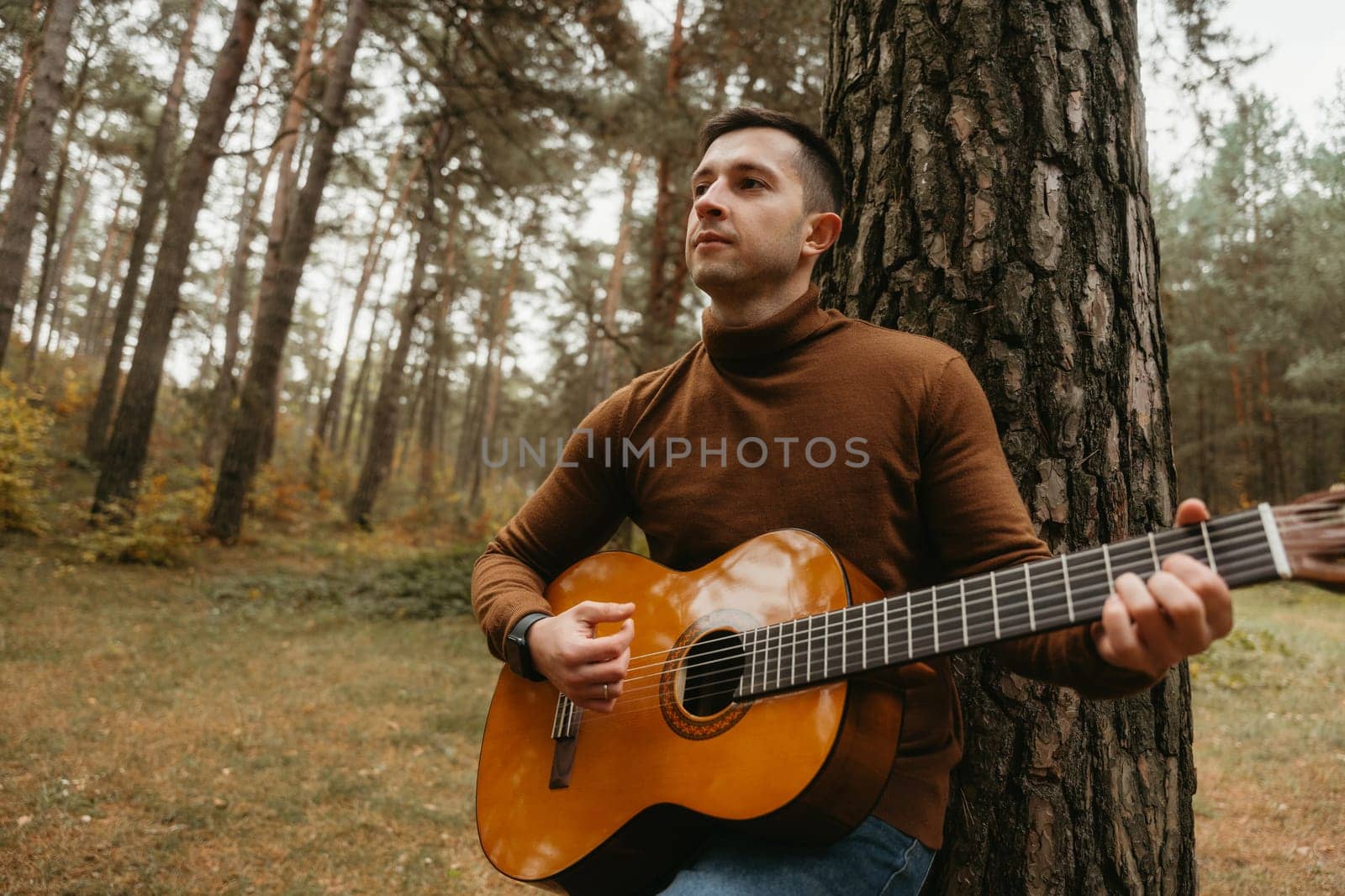Caucasian man plays guitar leaning on tree in woods, surrounded by nature