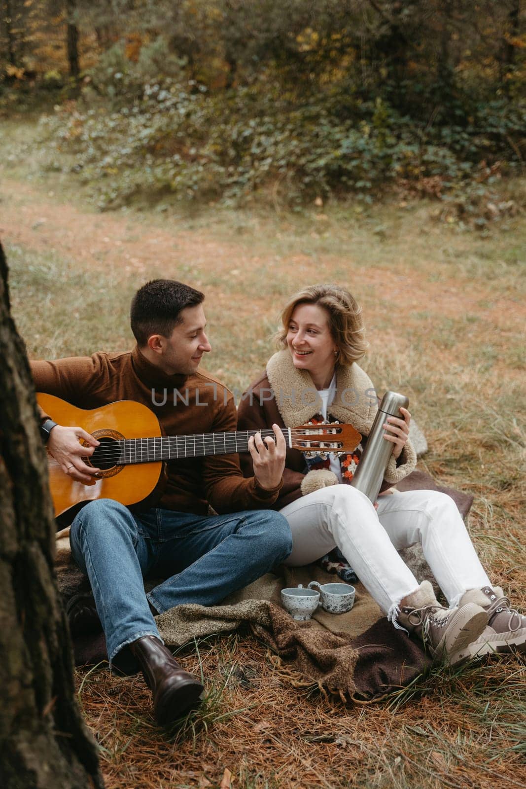 A man is playing guitar for happy woman, adult caucasian couple sitting on blanket in autumn park by Romvy