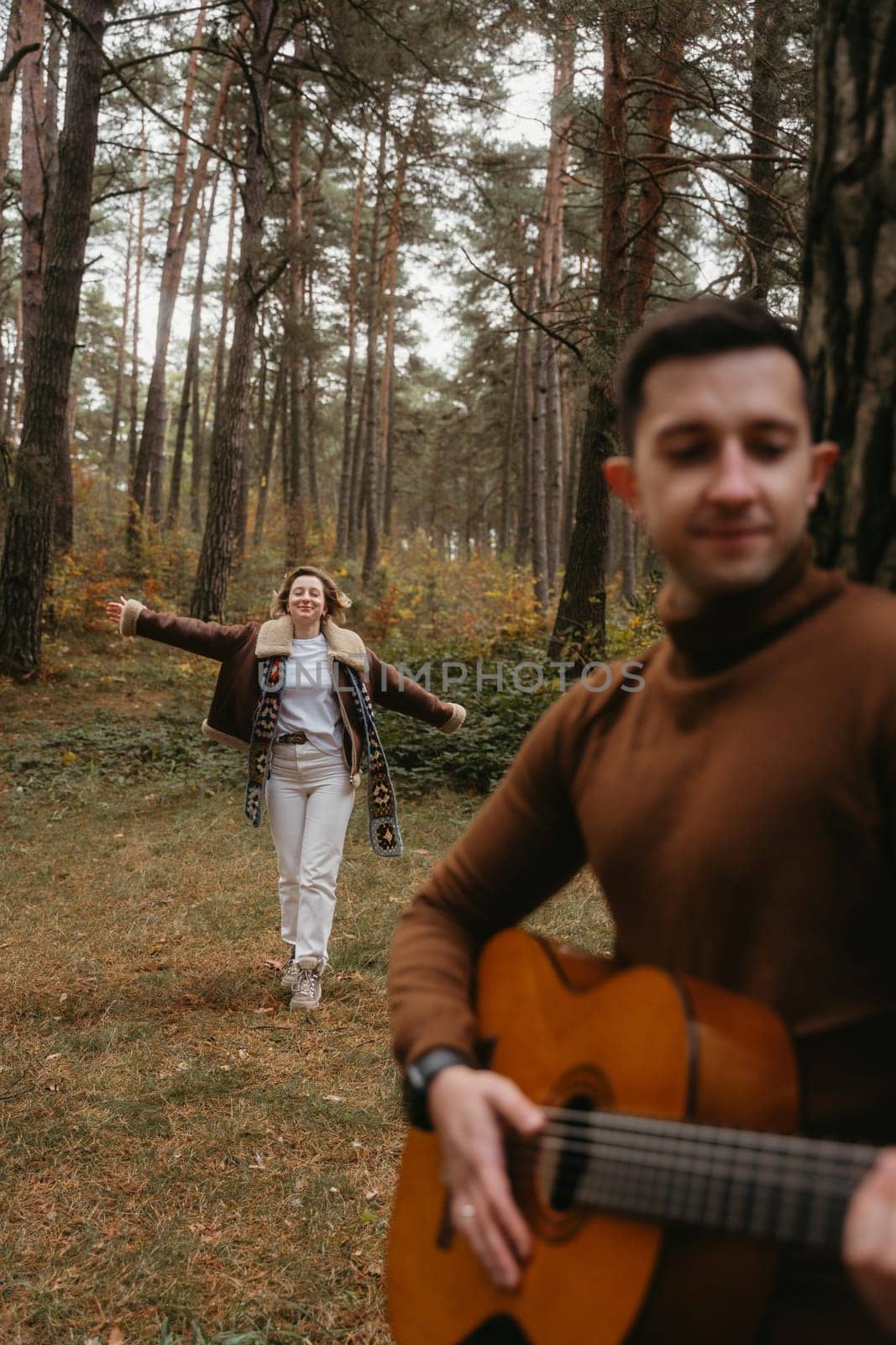 Man playing guitar outdoors in woods, a woman dancing behind him in the autumn park