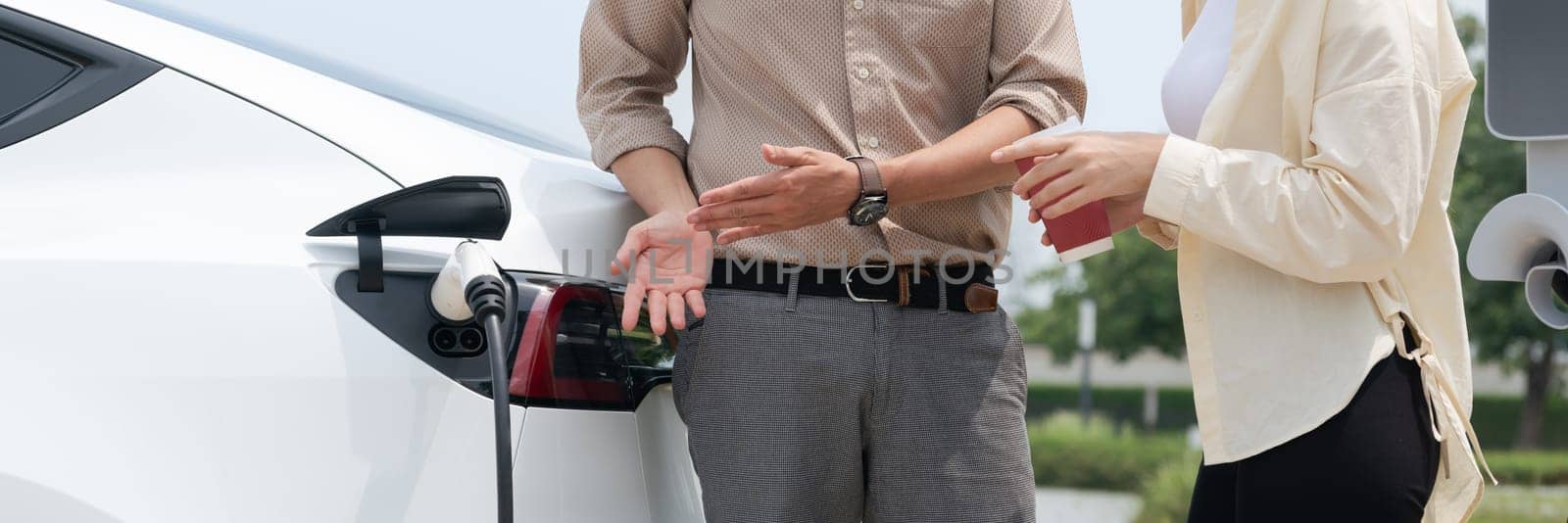 Young couple with coffee recharge EV car battery at charging station connect to electrical industrial power grid. Couple with shopping and travel using eco electric car lifestyle.Panorama Expedient