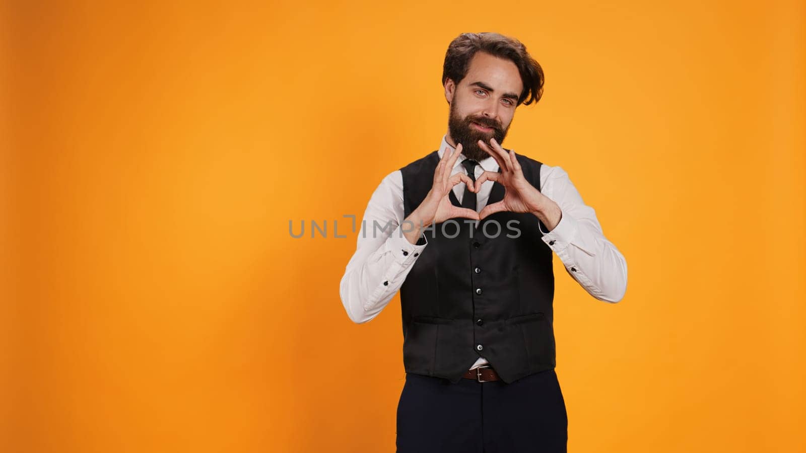 Butler showing heart shaped symbol in studio, working at five star restaurant and uncovering sincere feelings. Stylish waiter being romantic and presenting his love with sign, honest emotions.