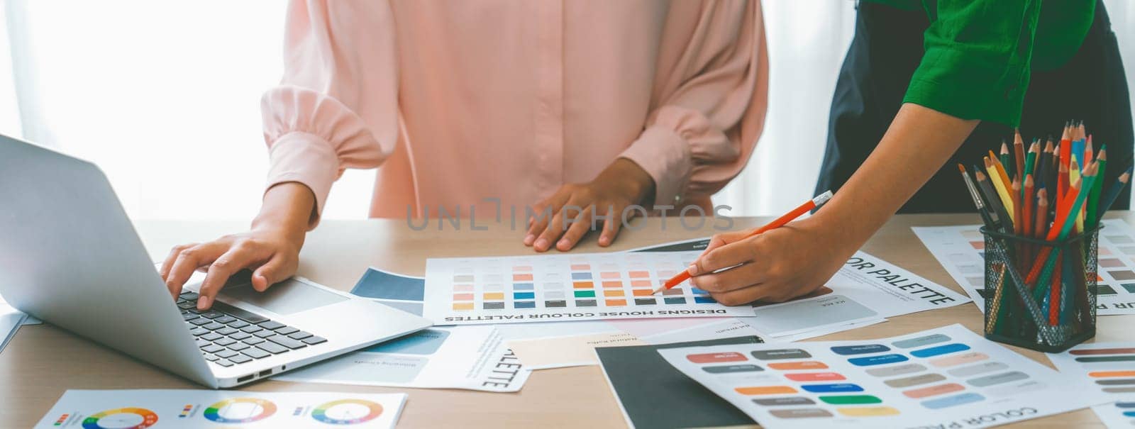 Cropped image of two female young beautiful graphic designer choose the color from color palate by using laptop on table with work tools and equipments scatter around at modern office. Variegated.